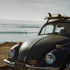 Image of VW bug at the beach with surfboards on top and surfers in the ocean.